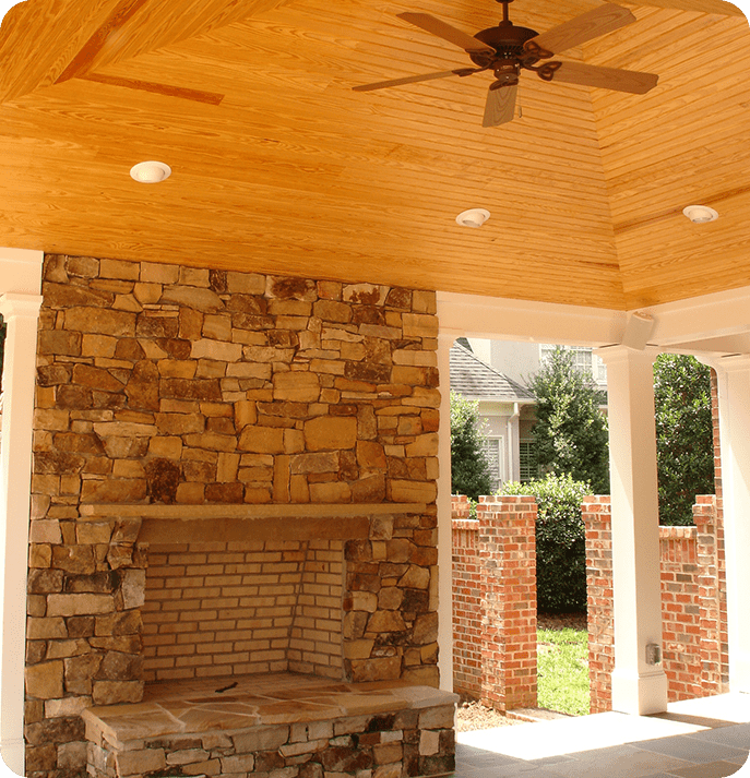 An outdoor patio area with a stone fireplace and a wooden ceiling fitted with a ceiling fan.